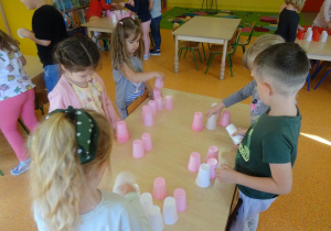 Pięcioro dzieci stoi przy stoliku i wspólnie próbuje ułożyć z różowych oraz białych plastikowych kubeczków uśmiech.